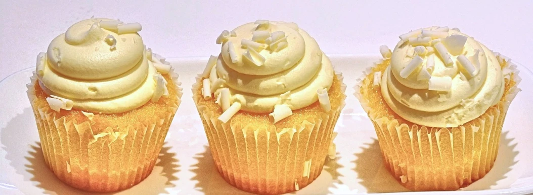 three cupcakes with white frosting on a white plate, by Teresa Fasolino, pixabay, light yellow, glittering and soft, it has lemon skin texture, blonde cream