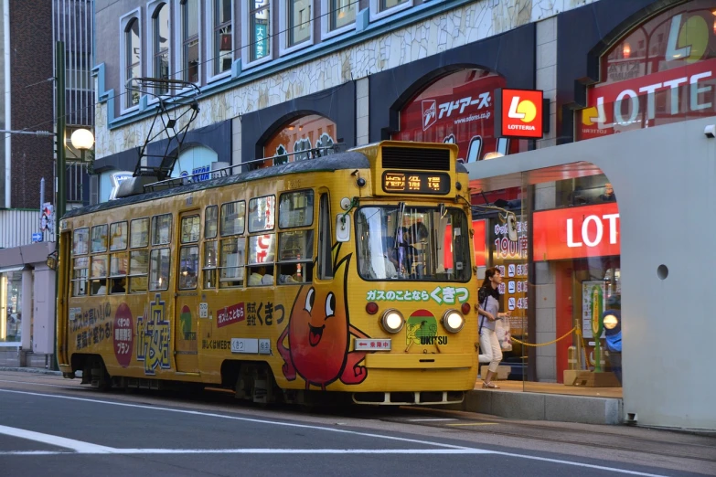 a yellow bus driving down a street next to a tall building, by Sengai, flickr, toyism, street tram, japanese mascot, pacman, summer morning