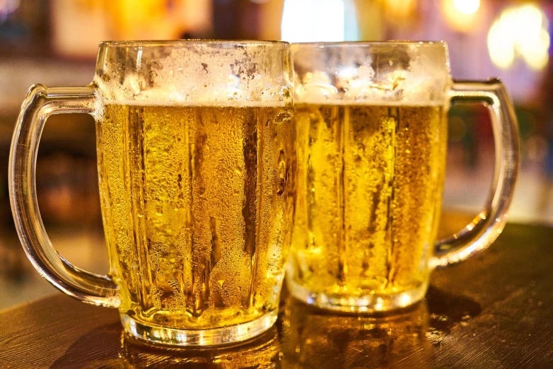 two glasses of beer sitting on top of a wooden table, by Adam Marczyński, pixabay, gold glasses, holding a mug of beer, golden glistening, zoomed in shots
