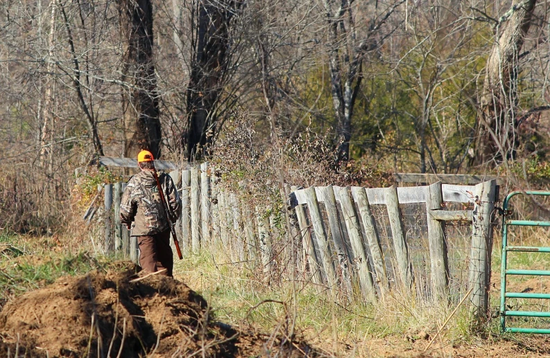 a man standing next to a pile of dirt, by Tom Carapic, fine art, carrying a scoped hunting rifle, fence line, walking away from camera, huntress