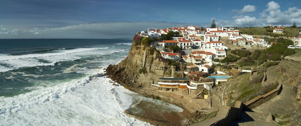 a group of houses sitting on top of a cliff next to the ocean, a picture, by Nadir Afonso, panoramic widescreen view, white buildings with red roofs, large waves hitting the cliff, beutiful!