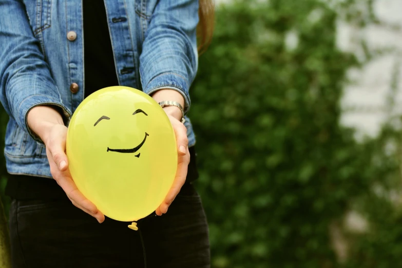 a woman holding a yellow balloon with a smiley face drawn on it, a picture, pexels, emotions closeup, small wide smile, a friendly wisp, background image