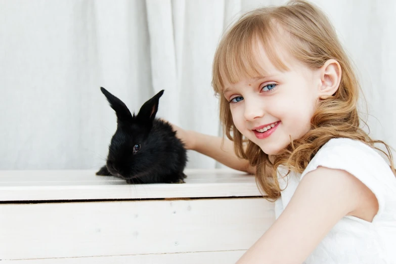 a little girl is petting a black rabbit, a stock photo, shutterstock, beautiful blue - eyes, on a white table, with a beautifull smile, two buddies sitting in a room
