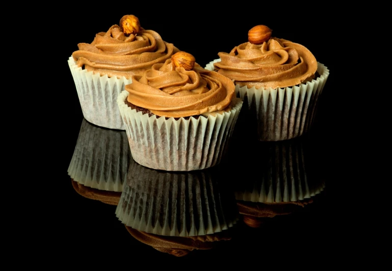 three cupcakes with chocolate frosting and nuts on top, by Etienne Delessert, against a deep black background, reflexions, high detail product photo