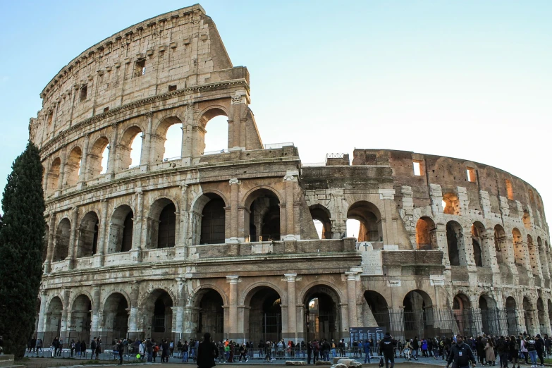 a group of people standing in front of a building, a picture, pexels contest winner, neoclassicism, colosseum, huge support buttresses, winter sun, buildings carved out of stone