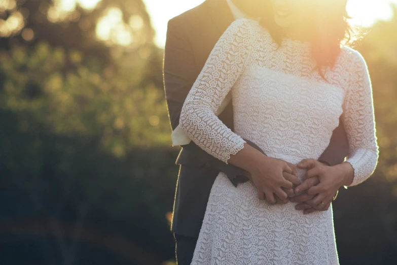 a man and a woman standing next to each other, by Joy Garnett, pexels, romanticism, simple cream dress, man grabbing a womans waist, with the sun shining on it, detailed lace dress