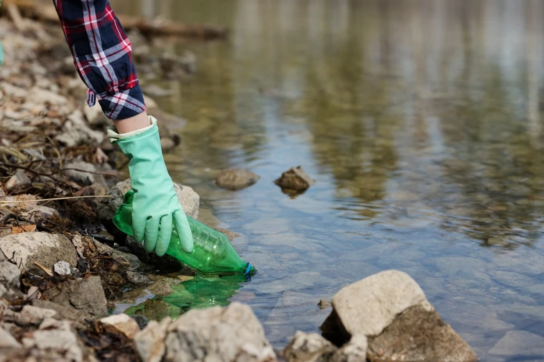 a person in green gloves standing next to a body of water, shutterstock, plasticien, creek, high res photo, holding a bottle, delete