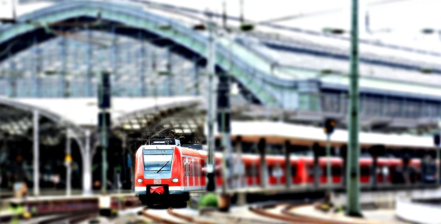 a red train traveling past a train station, a tilt shift photo, by Hans Schwarz, flickr, iphone photo, [ digital art ]!!, wallpaper - 1 0 2 4, munich