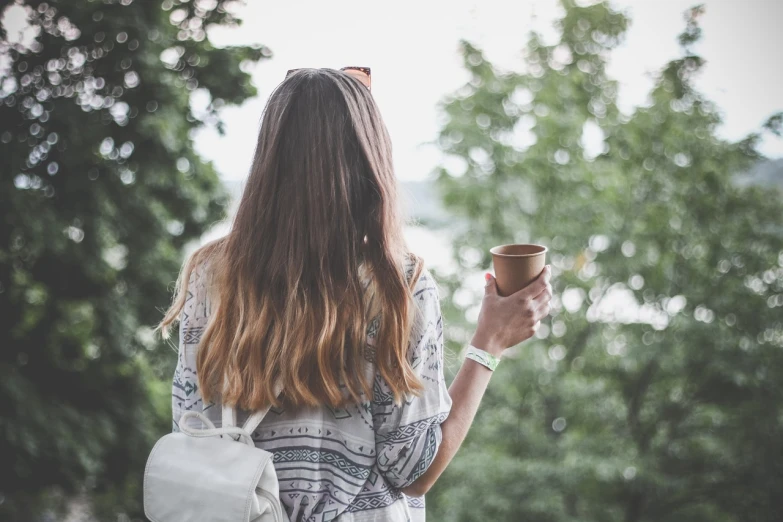a woman holding a cup of coffee in her hand, a picture, by Emma Andijewska, shutterstock, girl walking in forest, view from back, her hair is natural disheveled, with a backpack