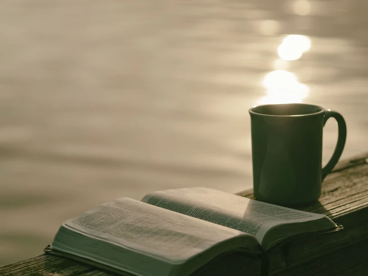 an open book sitting on top of a wooden bench next to a cup of coffee, a picture, by Romain brook, romanticism, glare on the water, mobile wallpaper, the lord and savior, in gentle green dawn light
