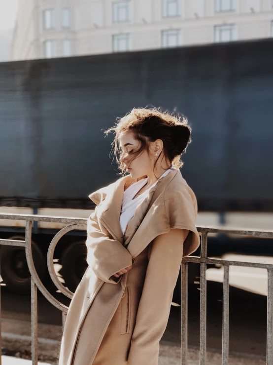 a woman leaning against a rail with a train in the background, a picture, by Emma Andijewska, trending on pexels, figuration libre, back of emma stone in beige coat, brunette woman, girl with messy bun hairstyle, fashion shoot 8k