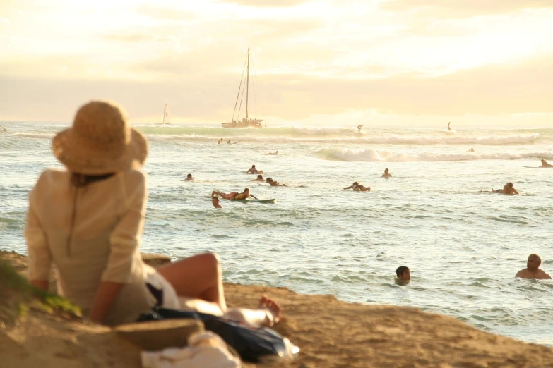 a woman sitting on top of a sandy beach next to the ocean, a picture, by Matt Stewart, waikiki beach, people watching, golden hues, people swimming