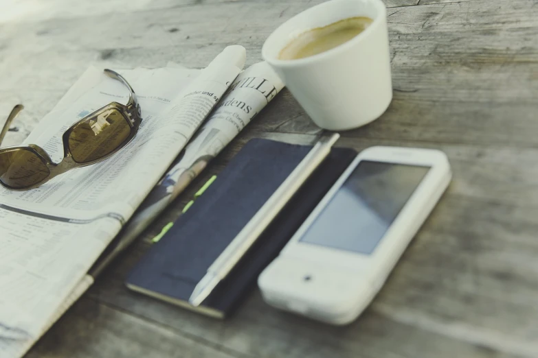 a cell phone sitting on top of a newspaper next to a cup of coffee, by Karl Buesgen, outdoor photo, vintage color, tools, very sharp photo