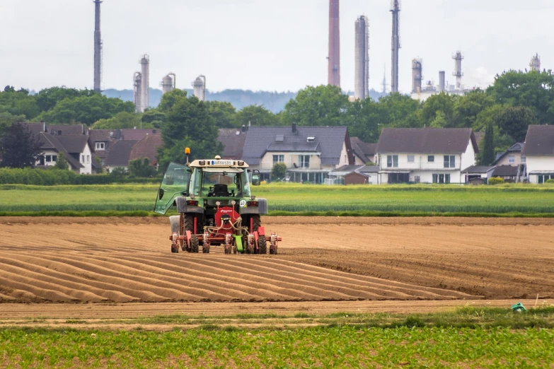 a tractor plowing a field in front of a factory, a picture, by Andries Stock, shutterstock, cultivator, chemical plant, reportage photo, thumbnail