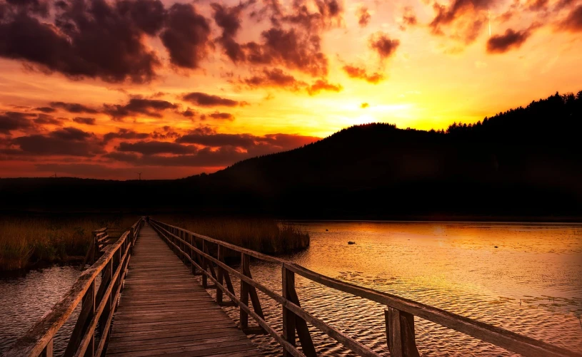 a wooden bridge over a body of water at sunset, by Yi Jaegwan, shutterstock, arrendajo in avila pinewood, moody sunset and dramatic sky, stock photo, on background red lake on fire