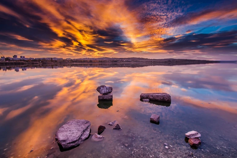 a group of rocks sitting on top of a body of water, a picture, by Gabor Szikszai, shutterstock, burning clouds, lake reflection, wide angle landscape shot, vivid colors!