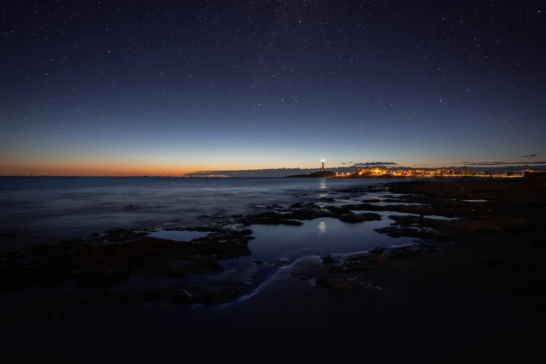 a beach at night with a lighthouse in the distance, a picture, by Carlo Martini, ethereal starlit city at sunset, south african coast, shiny city in the distance, stars reflecting on the water