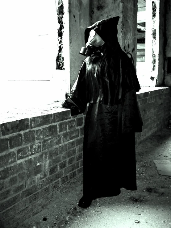 a man in a black coat leaning against a brick wall, a photo, inspired by Pietro Longhi, gothic art, she wear gasmask, covered in robes, monochrome:-2, grim reaper