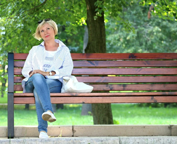 a woman sitting on top of a wooden bench, by Maksimilijan Vanka, pixabay, middle - age, white shirt and blue jeans, walking at the park, calmly conversing 8k