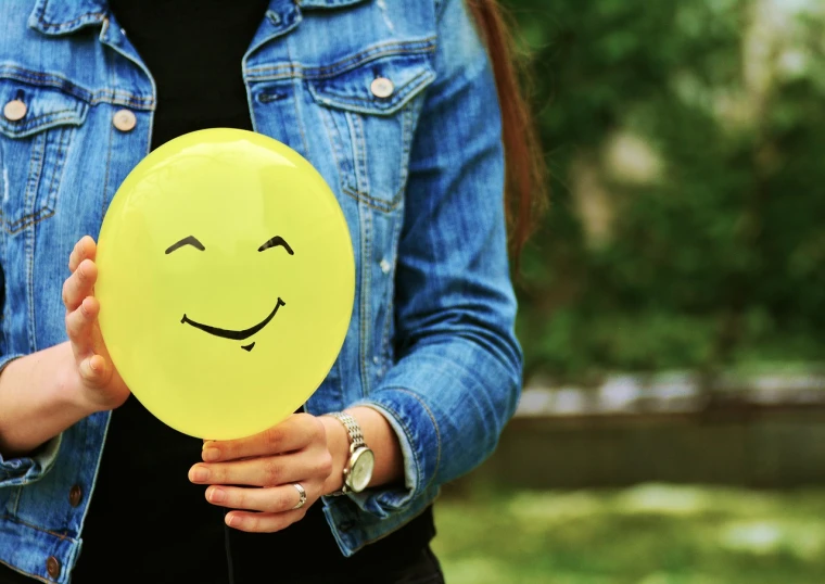 a woman holding a yellow balloon with a smiley face drawn on it, a picture, shutterstock, emotions closeup, stock photo