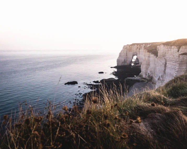 a man standing on top of a cliff next to the ocean, a picture, by Raphaël Collin, pexels, romanticism, white sweeping arches, northern france, early morning, wide long view