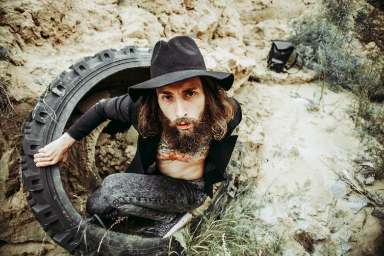 a man with a beard sitting inside of a tire, by Adam Szentpétery, hot summertime hippie, buried in sand, caracter with brown hat, young with long hair