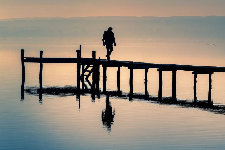 a man standing on a dock next to a body of water, by Zoran Mušič, shutterstock, people walking into the horizon, anthropomorphic silhouette, wooden bridge, soft reflections