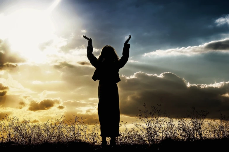 a woman standing on top of a grass covered field, a picture, by Eugeniusz Zak, pixabay, figuration libre, praying at the sun, arms raised, silhouetted, surrounded in clouds and light