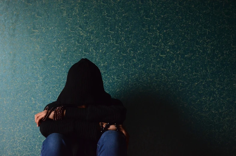 a person in a black hoodie sitting against a wall, pexels, antipodeans, grieving, hugging, sitting on a table, face covered