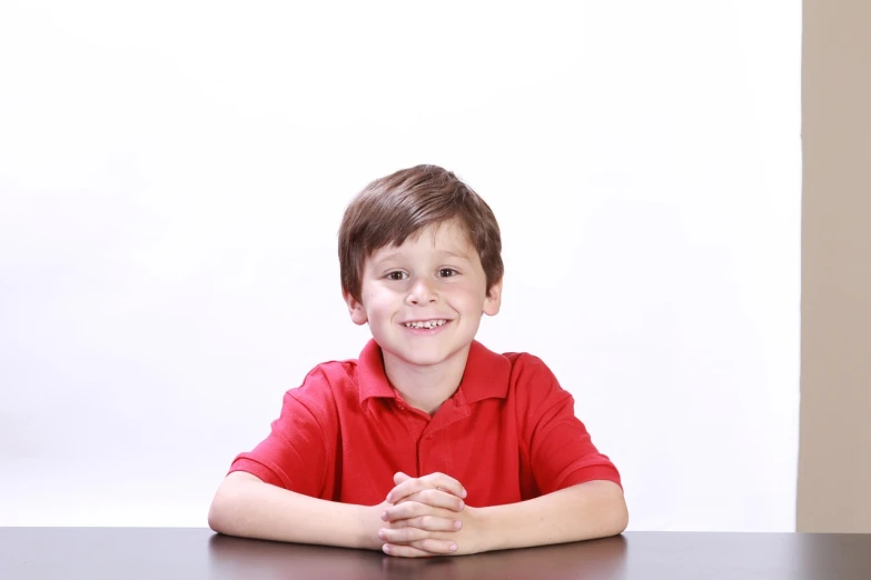 a young boy in a red shirt sitting at a table, a picture, istockphoto, in front of white back drop, zachary corzine, posing for a picture