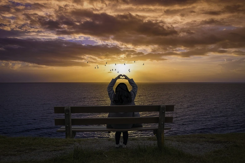 a woman sitting on a bench in front of the ocean, a picture, by Matt Stewart, pixabay contest winner, romanticism, hearts, sunset halo behind her head, love peace and unity, in love selfie