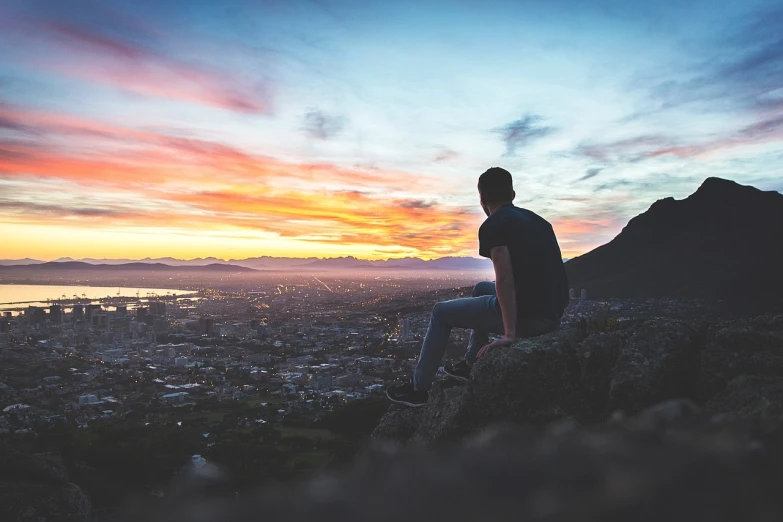 a man sitting on top of a rock overlooking a city, by Jakob Gauermann, pexels, colorful sunset, cape, teenage boy, istockphoto