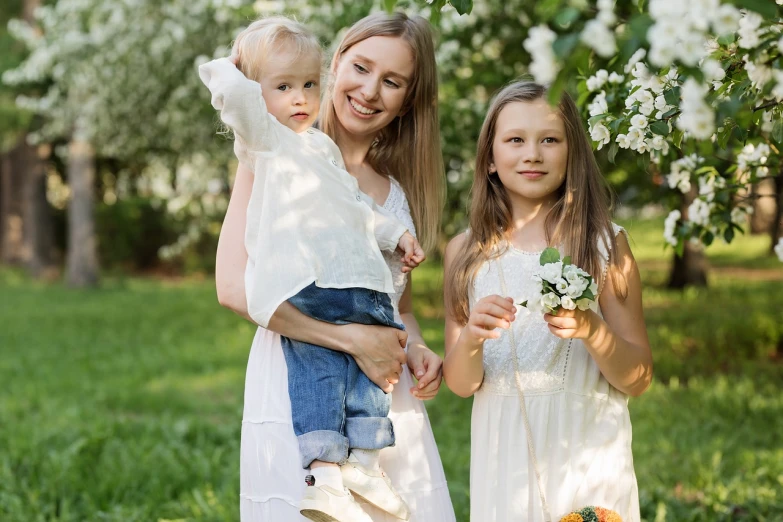 a woman holding a baby and two young girls, by Maksimilijan Vanka, pexels, white blossoms, with fruit trees, avatar image, 7 0 mm photo