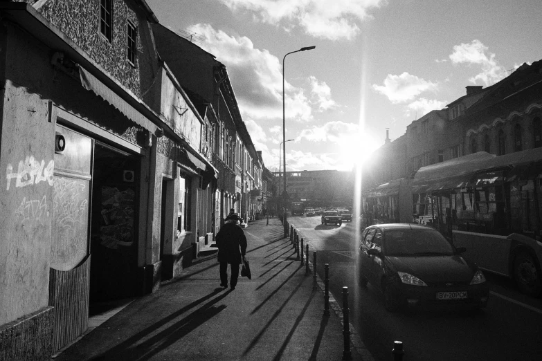 a black and white photo of a person walking down a street, a black and white photo, by Tamas Galambos, winter sun, small town, nice afternoon lighting, people walking around