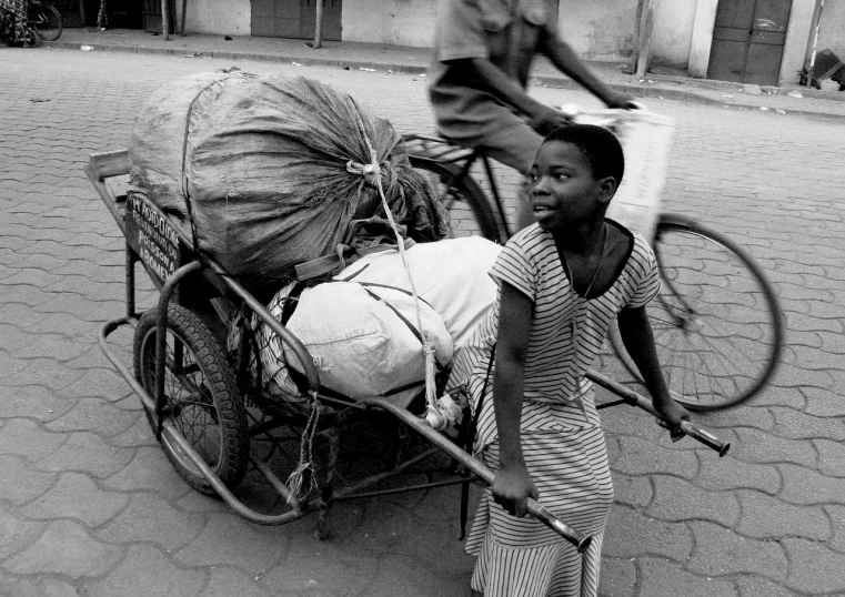 a black and white photo of a boy pushing a bike with a sack on the back, by Albert Welti, flickr, light skinned african young girl, cart, shot with a arriflex 35 ii, 1996)