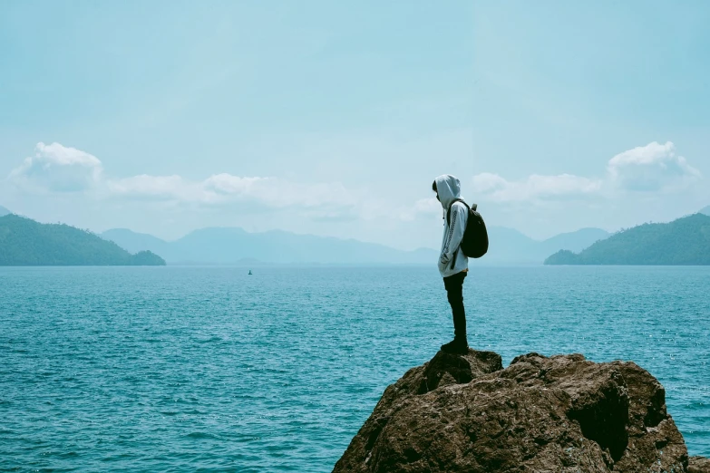 a person standing on top of a rock near the ocean, a picture, minimalism, a man wearing a backpack, wearing a blue hoodie, teenage boy, with his back turned