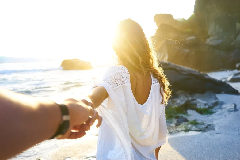 a woman holding the hand of a man on a beach, a picture, into bright light, flowy, back towards camera, wearing white clothes