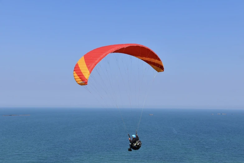 a person parasailing over the ocean on a sunny day, a picture, by Edward Corbett, shutterstock, red and yellow scheme, ornithopter, canopy, cliffside