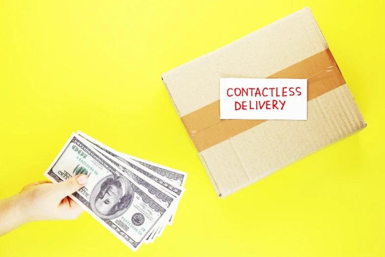 a hand holding a stack of money next to a cardboard box with contactless delivery written on it, a stock photo, by Allen Jones, red contact lenses, on a yellow canva, ny, banner