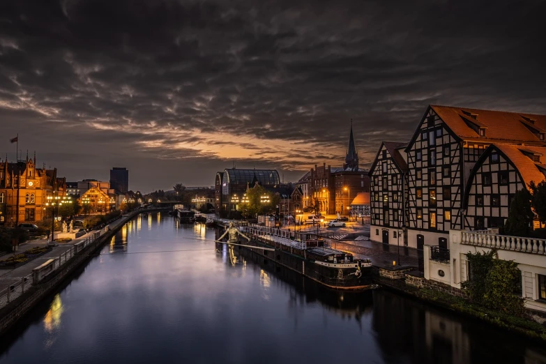 a river running through a city next to tall buildings, a photo, by Erik Pevernagie, baroque, moody sunset and dramatic sky, in legnica, docks, dutch houses along a river