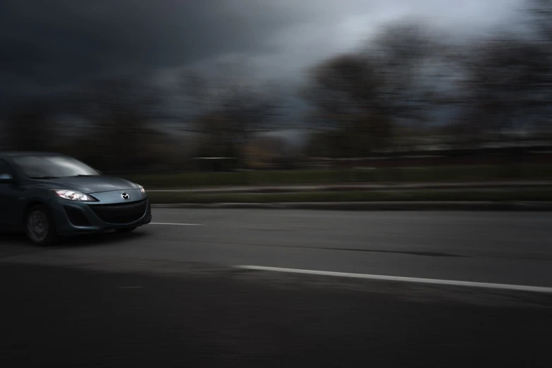a car driving down a road under a cloudy sky, a picture, by Andrew Domachowski, background ( dark _ smokiness ), panning shot, wide body, underexposed grey