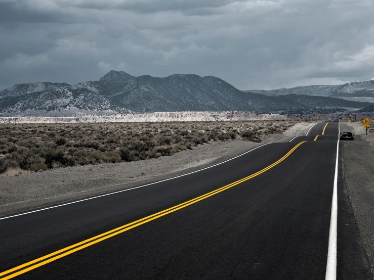 a car driving down a road with mountains in the background, a picture, by Andrew Domachowski, photorealism, surreal black and yellow, kodachrome photograph, dark overcast weather, desert highway