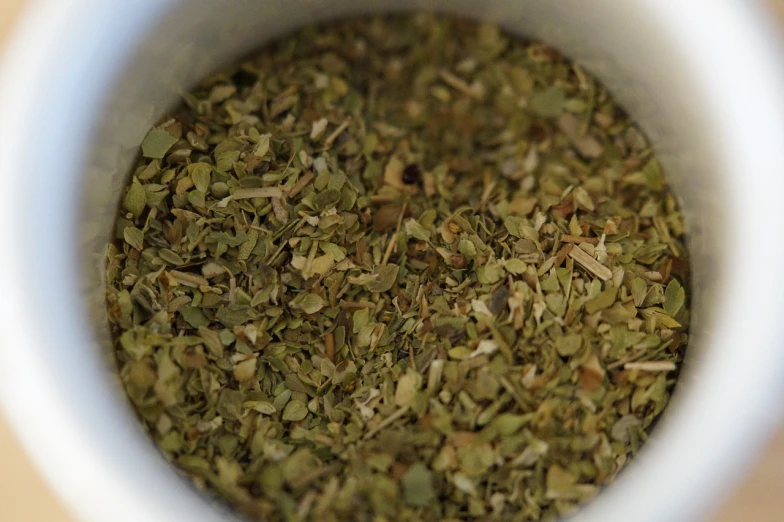 a white cup filled with dried herbs on top of a table, a picture, by William Berra, hurufiyya, chewing tobacco, looks a blend of grimes, upper body close up, olive thigh skin