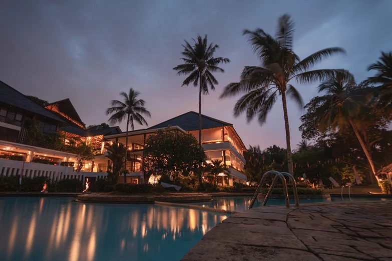 a swimming pool surrounded by palm trees at night, a stock photo, by Bernardino Mei, shutterstock, hotel, slight overcast lighting, during sunset, cinematic front shot