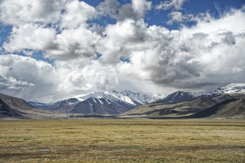 a large open field with mountains in the background, by Muggur, snow capped mountains, dramatic clouds and atmosphere, empty remote wilderness, dessert