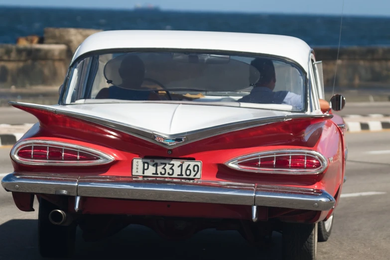 a red and white car driving down a street next to the ocean, classic cars, detail shot, cuban revolution, back shark fin