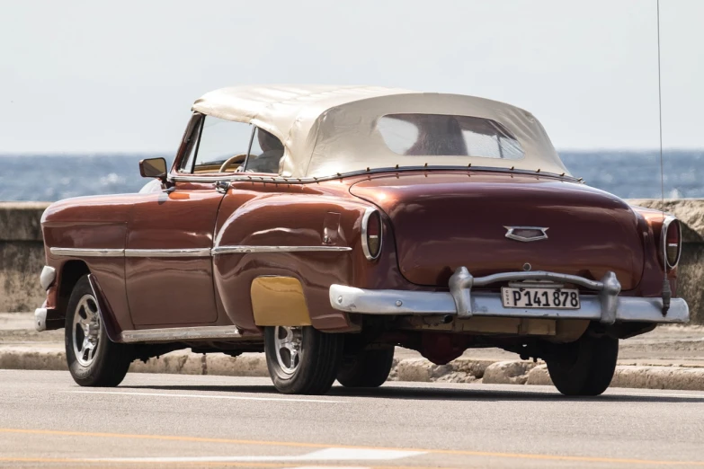 an old car driving down a road next to the ocean, modernism, soft top, less detailing, cuban setting, tail fin