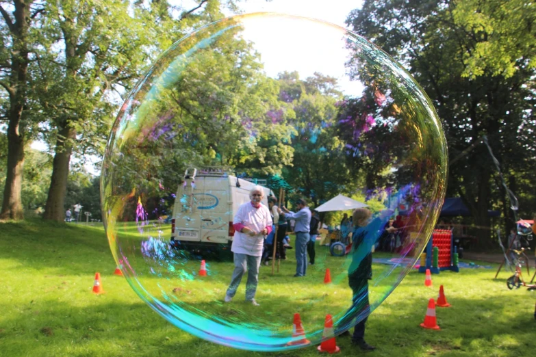 a group of people standing on top of a lush green field, iridescent soapy bubbles, community celebration, magic bubble barrier, time travelers appear in a park