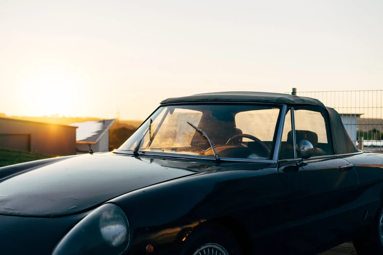 a man sitting in the driver's seat of a black car, unsplash, romanticism, soft top roof raised, sun at dawn, alfa romeo project car, nostalgic vibes