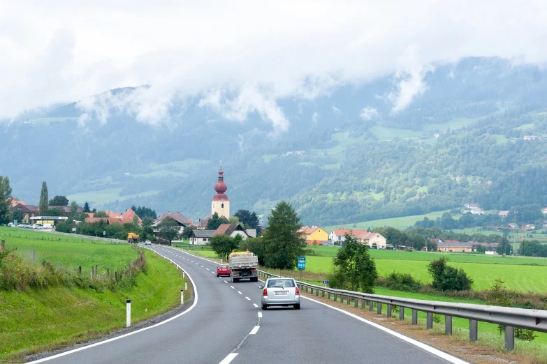 a car driving down a road next to a lush green hillside, a picture, by Franz Hegi, shutterstock, church in the background, austria, nice slight overcast weather, stock photo